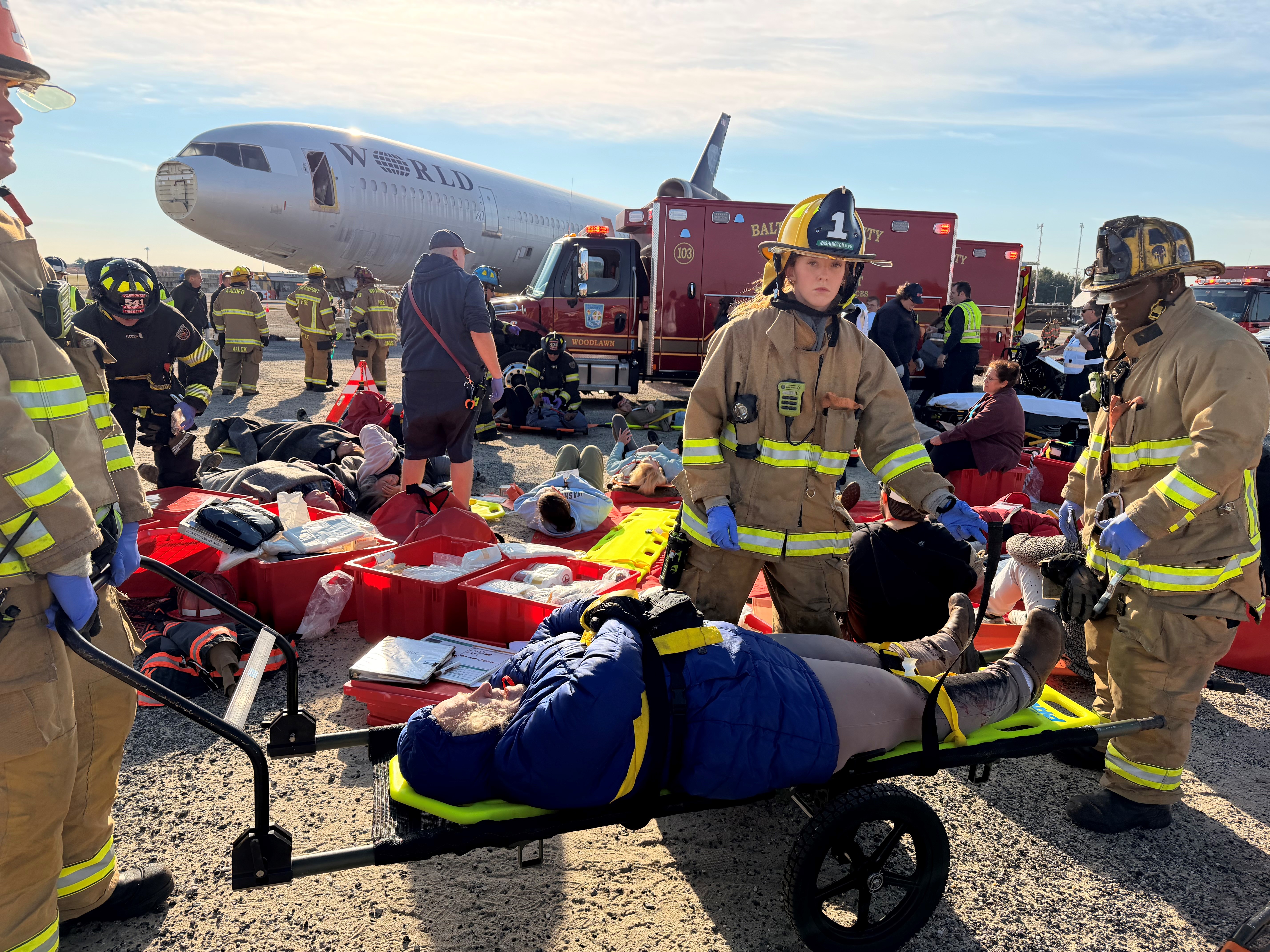 Emergency Medicine residents dress in fire-protectant gear to support patient on a gurney outdoors at an airport terminal.